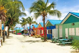 Colourful beachside cottages line a sandy path bordered by palm trees in a vibrant Caribbean setting | MSC Cruises
