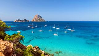 A serene view of a turquoise sea with sailboats near rocky islands under a clear blue sky | MSC Cruises