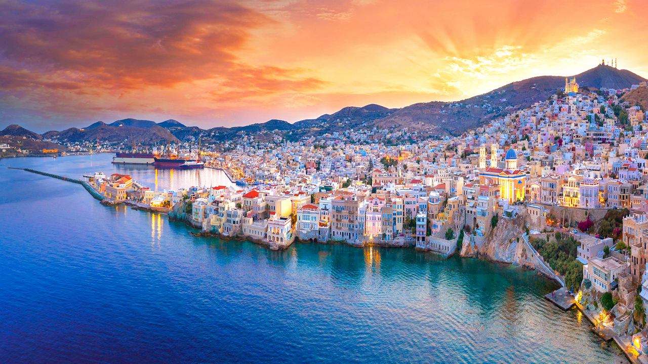 Ein atemberaubender Blick auf eine Küstenstadt in Griechenland mit lebhaften Gebäuden und ruhigen Gewässern bei Sonnenuntergang | MSC Cruises