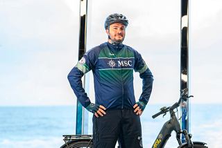A bike tour leader stands with a bicycle near the sea and the MSC Euribia ship in the background | MSC Cruises