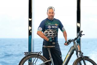 Happy MSC Euribia crew member with bike by the sea and ship in the background | MSC Cruises