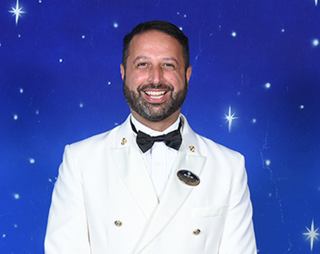 A smiling captain in a formal white uniform poses confidently against a starry blue background | MSC Cruises