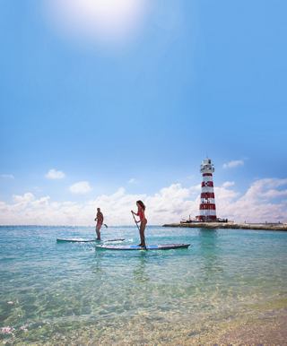 Couple paddleboarding by a lighthouse in clear blue water beneath the sun | MSC Cruises