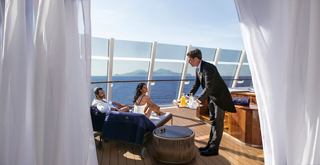 A butler serves drinks to a couple relaxing on a balcony overlooking the sea and mountains | MSC Cruises