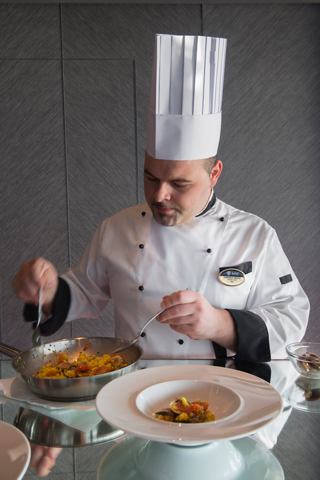 A chef in a white uniform prepares a gourmet dish in a refined dining setting | MSC Cruises