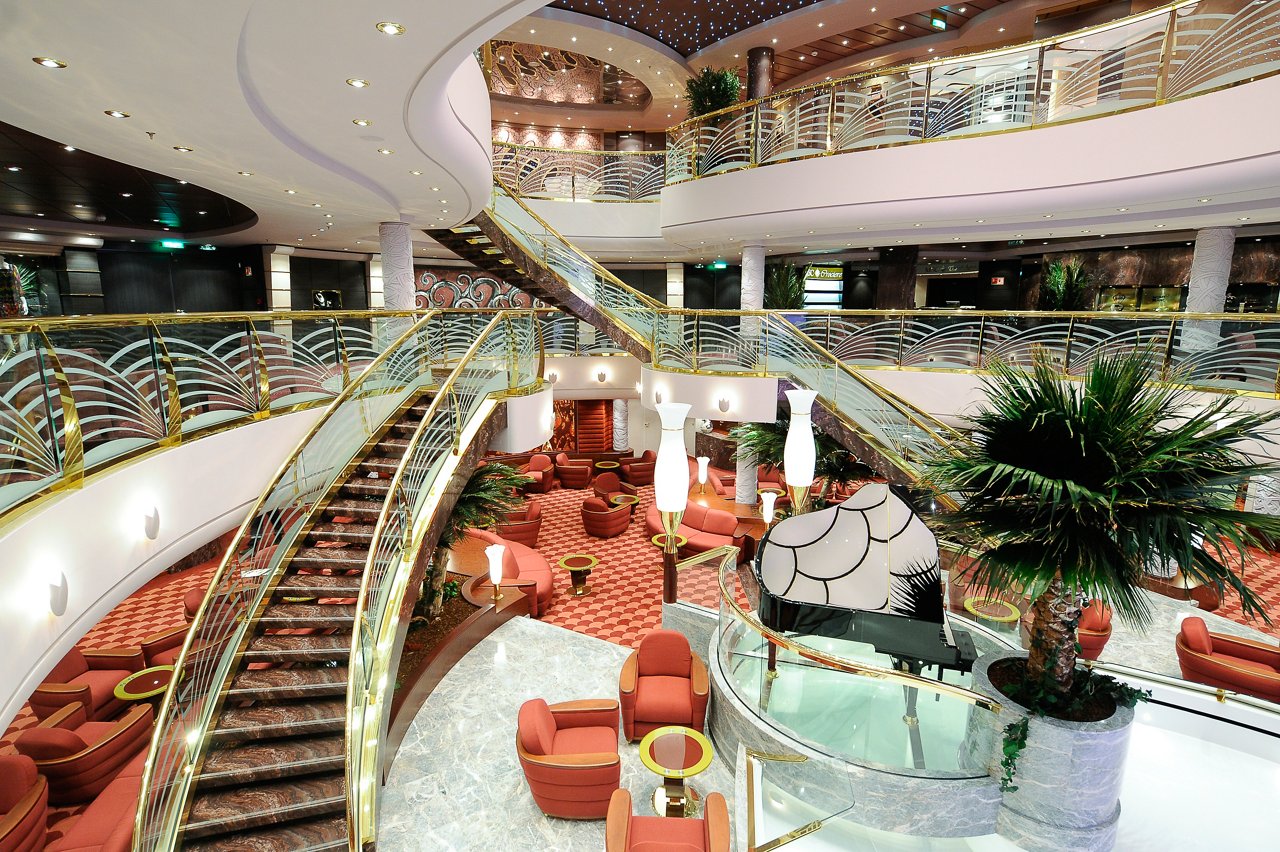 A spacious reception area features elegant seating, a grand staircase, and lush greenery, welcoming guests aboard MSC Magnifica | MSC Cruises