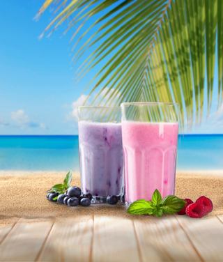 Two vibrant smoothies in tall glasses, one pink and one purple, sit on a sandy beach table with tropical scenery | MSC Cruises