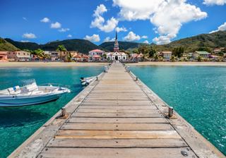 A wooden jetty extends over clear turquoise water, leading to a charming coastal village with a church in the background | MSC Cruises