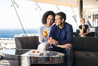 A couple enjoys cocktails and snacks together in the Sky Lounge, smiling with the ocean view in the background | MSC Cruises