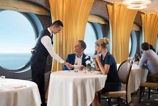 A waiter serves dessert to a couple enjoying a meal by a large porthole on MSC Meraviglia | MSC Cruises