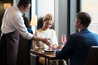 A couple enjoys a meal at a specialty restaurant, with a waiter serving them drinks on MSC Meraviglia | MSC Cruises