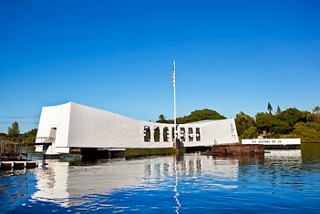 A modern memorial building reflecting in the calm waters, surrounded by lush vegetation and a clear blue sky | MSC Cruises
