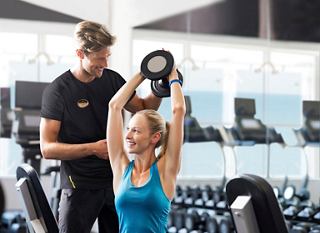 Un entrenador ayuda a una mujer con un ejercicio de levantamiento de pesas en un entorno de gimnasio moderno | MSC Cruceros