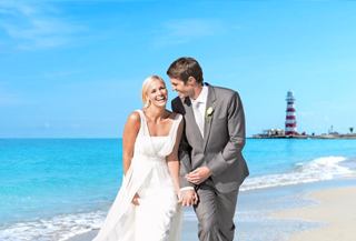 Ein glückliches Paar umarmt sich am Strand mit einem Leuchtturm und ruhigem Meer im Hintergrund | MSC Cruises