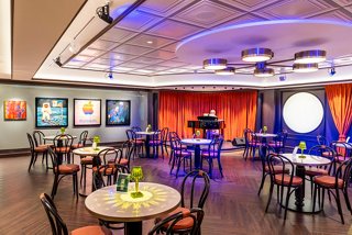 A stylish lounge area features tables and chairs, with vibrant artwork and a piano creating a welcoming atmosphere | MSC Cruises