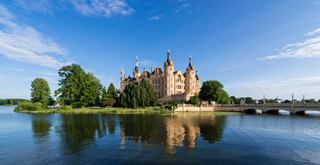 Ein majestätisches Schloss, das sich in einem ruhigen See spiegelt, umgeben von üppigen Bäumen und einem klaren blauen Himmel | MSC Cruises