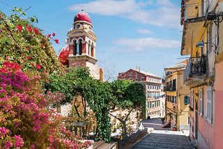 A vibrant street in Corfu, featuring blooming flowers and a charming tower against a clear blue sky | MSC Cruises