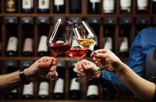 Two hands clinking glasses of red and white wine in a warm toast against a backdrop of wine bottles | MSC Cruises