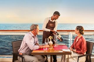 A couple enjoys a meal at a seaside table as a crew member serves them with a scenic ocean backdrop | MSC Cruises