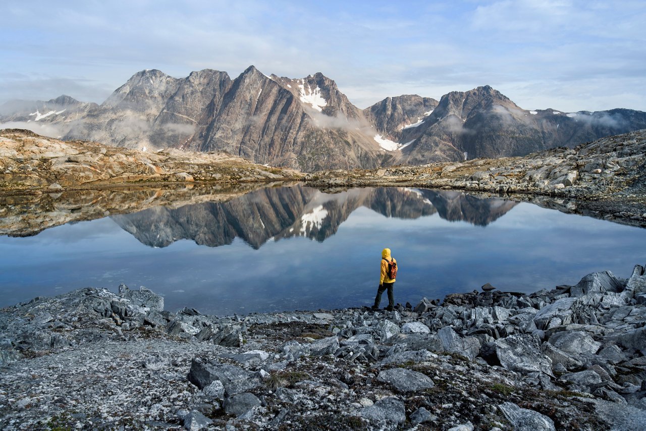 A person stands by a serene lake surrounded by majestic mountains in Greenland's stunning landscape | MSC Cruises
