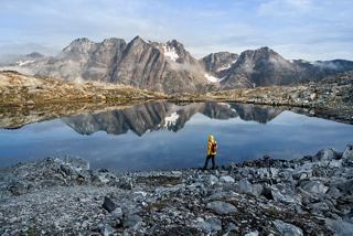 A person stands by a serene lake surrounded by majestic mountains in Greenland's stunning landscape | MSC Cruises