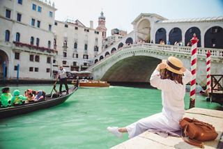 Une femme portant un chapeau est assise au bord du canal à Venise, admirant la vue sur le pont du Rialto | MSC Croisières