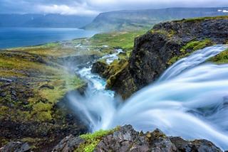 A breathtaking view of Isafjordur's cascading waterfall amidst lush greenery and rocky cliffs by the coast | MSC Cruises