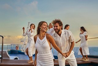 A couple enjoys a lively moment together on deck during a white party, surrounded by friends dancing in the sunset | MSC Cruises