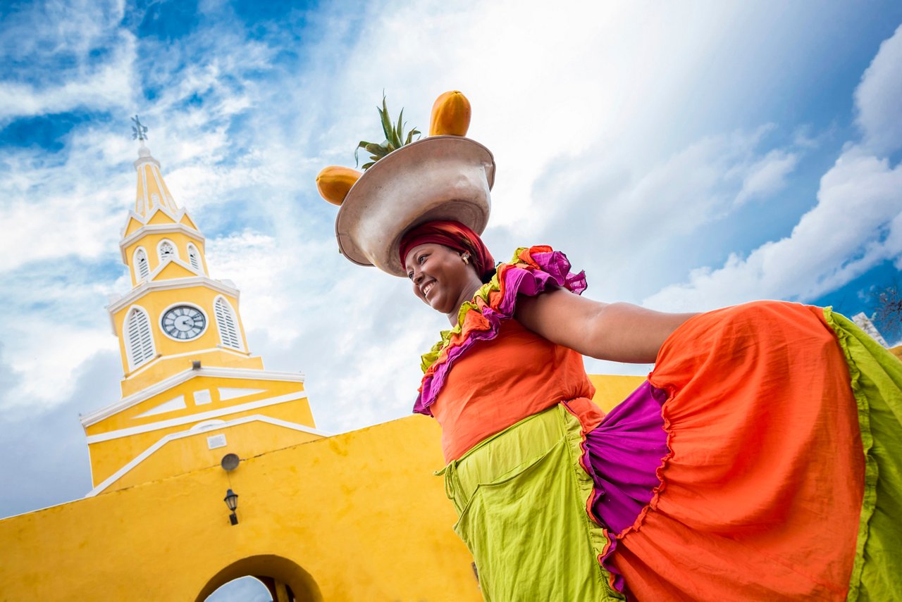 A woman in vibrant traditional attire poses near a colourful building in Cartagena under a bright blue sky | MSC Cruises
