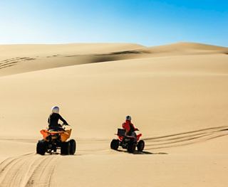 Dos personas montan ATV a través de dunas de arena doradas bajo un cielo azul claro en el paisaje desértico de Namibia | MSC Cruceros