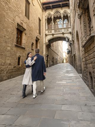 A young couple walks arm in arm down a narrow stone street between tall historic buildings. Ornate Gothic-style architecture with an elevated bridge connects the structures above the alley. The scene appears quiet and peaceful, with soft daylight illuminating the textured stone walls and pavement. Their long coats and relaxed posture add a sense of romance and leisurely exploration in an old European city.