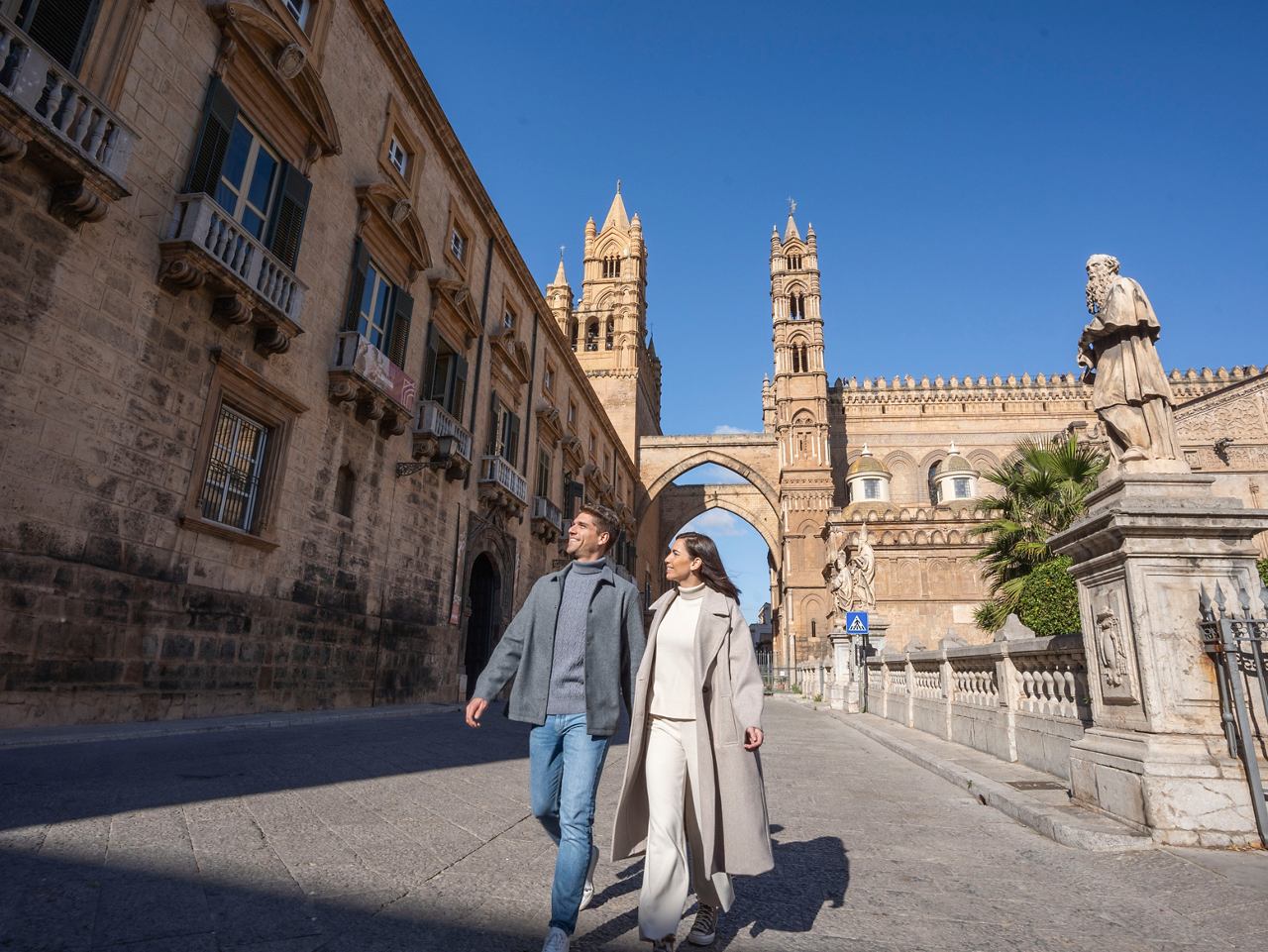 A couple strolls together in a historic square with a cathedral and arch in the background under a clear blue sky | MSC Cruises
