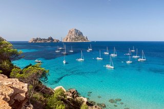 A scenic view of Cala Hort with Es Vedra in the background and boats on the turquoise sea under a clear sky | MSC Cruises