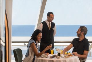 A couple enjoys breakfast with a waiter in the MSC Yacht Club restaurant, overlooking the sea through a glass wall | MSC Cruises