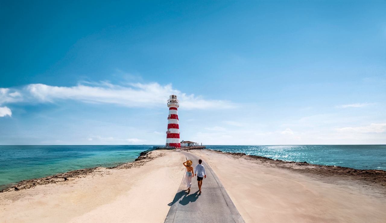A vibrant lighthouse stands at the end of a long pier, surrounded by sparkling blue waters and clear skies | MSC Cruises