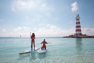 Una coppia si diverte a fare stand-up paddleboarding in acque cristalline vicino a un faro presso Ocean Cay MSC Marine Reserve | MSC Crociere