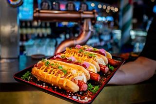 A hand holds a tray with three delicious hot dogs in a lively dining setting | MSC Cruises