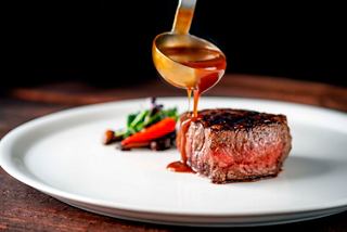 A gourmet steak garnished with fresh vegetables and a rich sauce being drizzled on top | MSC Cruises