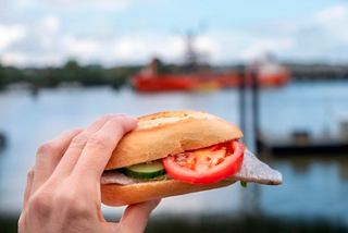 Hand holding a herring burger with tomato and cucumber against the backdrop of Hamburg harbor | MSC Cruises