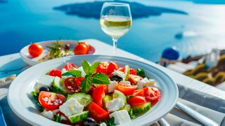 A vibrant salad with feta and tomatoes is accompanied by a glass of wine, set against the stunning Mediterranean backdrop | MSC Cruises