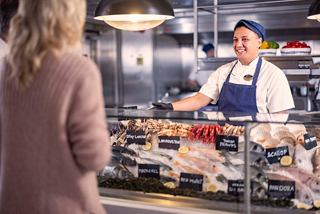 Un miembro amable de la tripulación sirve mariscos frescos en La Pescaderia mientras un huésped disfruta de la vibrante atmósfera | MSC Cruceros