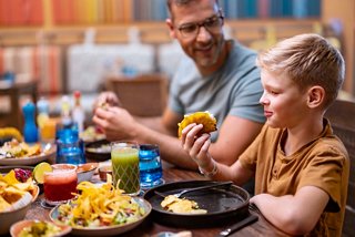 Ein Vater und sein Sohn genießen gemeinsam eine lebhafte Mahlzeit an einem bunten Restauranttisch voller Essen und Getränke | MSC Cruises