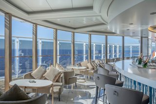 A stylish champagne bar with a glass wall offering stunning views, featuring tables and sofas for guests to relax | MSC Cruises