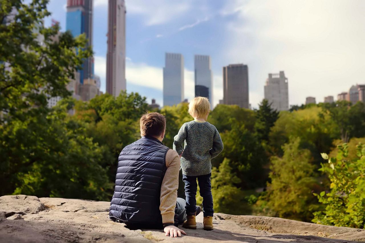 Ein Vater und sein Sohn entspannen sich auf einem Stein im Central Park mit Blick auf die Wolkenkratzer von New York und das üppige Grün | MSC Cruises