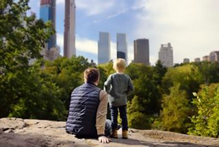 Un padre y su hijo se relajan sobre una roca en Central Park, con vistas a los rascacielos de Nueva York y la vegetación exuberante | MSC Cruceros