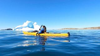 A woman paddles a bright yellow kayak in the serene sea of Greenland, surrounded by floating ice and clear skies | MSC Cruises
