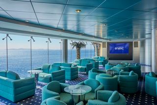 A vibrant lounge area with turquoise seating and a large screen, offering stunning sea views aboard MSC Lirica | MSC Cruises