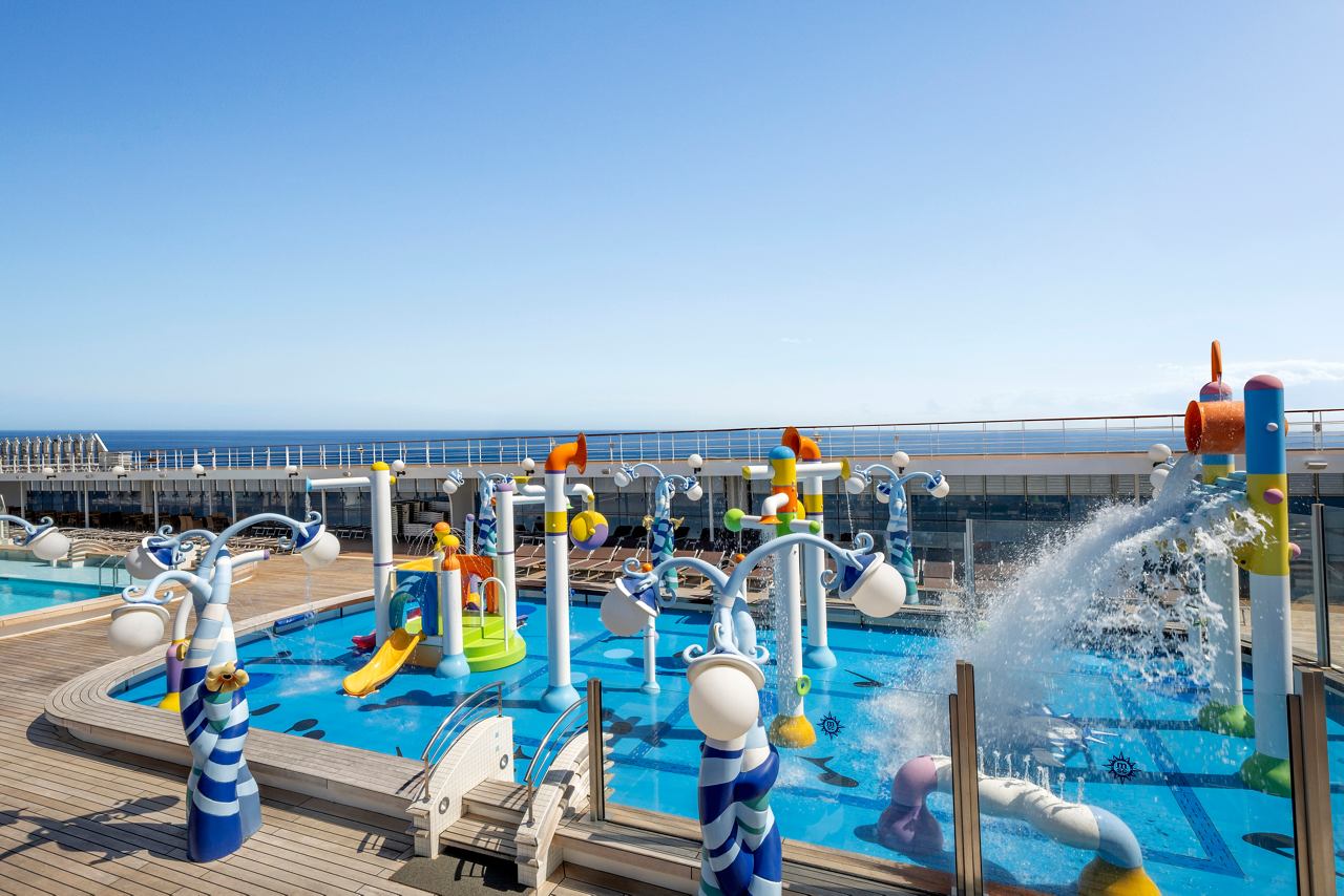 Ein lebendiger Wasserspielbereich mit bunten Elementen, Sonnenliegen und einem atemberaubenden Meerblick auf der MSC Lirica | MSC Cruises