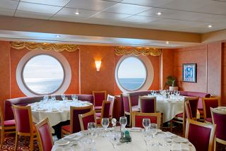 A warm and inviting dining area with round tables and elegant decor, featuring large portholes overlooking the sea | MSC Cruises