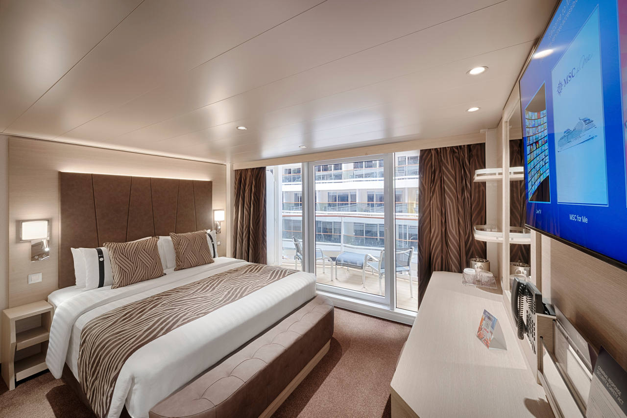 A luxurious Deluxe Balcony cabin featuring a double bed, vanity area, and outdoor furniture with a promenade view | MSC Cruises